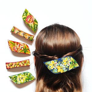 Peut inclure: Plusieurs barrettes à cheveux peintes à la main avec des motifs floraux. Les barrettes, de formes et de couleurs variées, comprennent du vert, du rouge et du bleu, ornées de fleurs jaunes, roses et blanches. Une barrette est dans les cheveux d'une femme.