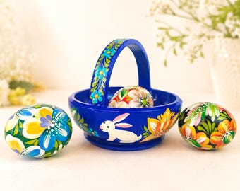 May include: A bright blue wooden basket with a handle, featuring hand-painted white rabbit and floral motifs. The basket contains one decorative egg, while two additional hand-painted eggs with colorful floral patterns sit on either side.
