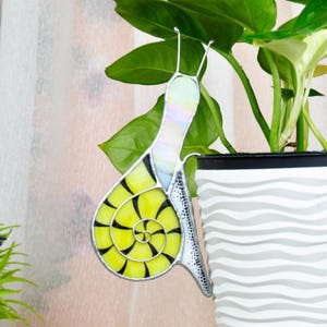 May include: A stained glass snail decoration with a yellow shell and black spiral design. The snail's body has iridescent and clear glass. The snail is hanging near a green plant in a white and gray striped pot.
