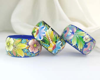 May include: Three hand-painted wooden bangles. The bangles are decorated with colorful floral designs in shades of blue, yellow, pink, and green. The background is a soft white fabric.