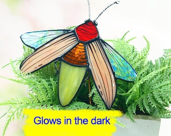May include: A stained glass insect sculpture with a red head, orange and tan body, and green and blue wings. The text "Glows in the dark" is visible on a yellow background. The sculpture is placed in a pot of green plants.