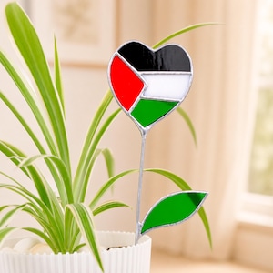 May include: A stained glass plant stake featuring a heart-shaped top with the colours of the Palestinian flag in black, white, green, and red. The piece includes a long metal stem and a single green stained glass leaf attached to the side.