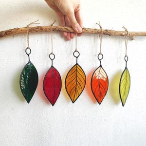 May include: Five stained glass leaf ornaments in green, red, yellow, orange, and light green, suspended from a wooden branch. Each leaf has a black metal outline and is hung by twine.