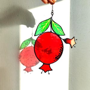 May include: A stained glass pomegranate suncatcher with a red fruit, green leaves, and a small flower. The fruit has black speckles and is outlined in black. It hangs from a chain and casts a shadow on a white wall.