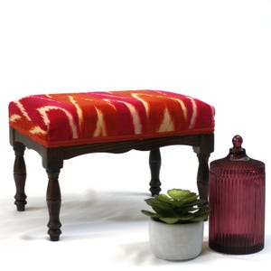May include: A wooden footstool with four turned legs and a red, orange, and white ikat fabric top.
