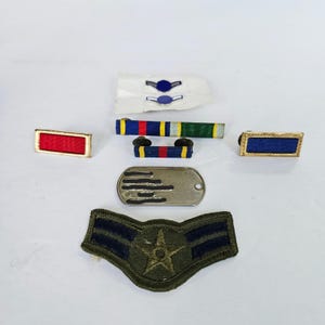 May include: A collection of military insignia including a red ribbon bar, a blue ribbon bar, a multi-coloured ribbon bar, a smaller ribbon bar with red and blue stripes, a silver-toned metal identification tag, and an olive green chevron patch.