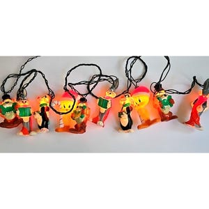May include: A string of vintage Christmas lights featuring cartoon character figures. The figures are brightly colored, with a warm orange glow emanating from within. Each figure is attached to a black wire.