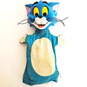 May include: A blue Tom Cat hand puppet with a smiling face, yellow eyes, and black whiskers. The puppet's body is blue corduroy with a cream-colored oval on the front and cream-colored hands.