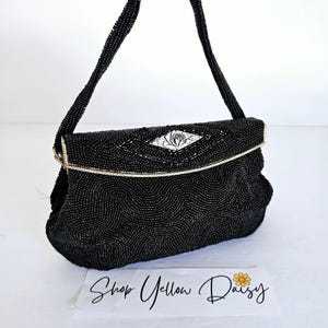 Puede incluir: Un bolso de mano negro con cuentas con un cierre y una correa de color plateado. El bolso presenta un adorno en forma de diamante con un diseño floral. La bolsa está sobre una superficie blanca con el texto "Shop Yellow Daisy".