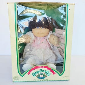 May include: A 1984 Cabbage Patch Kids doll with dark brown yarn hair and blue eyes, wearing a pink and white patterned dress. The doll is inside its original rectangular cardboard box with a clear plastic window and includes a birth certificate.