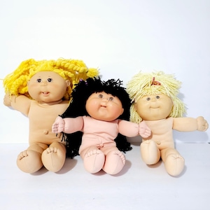Może przedstawiać: Trzy lalki Cabbage Patch Kids z różnymi kolorami włosów: żółtym, czarnym i blond. Lalki mają miękkie, materiałowe ciała i siedzą z wyciągniętymi ramionami. Lalki mają klasyczny, vintage wygląd.