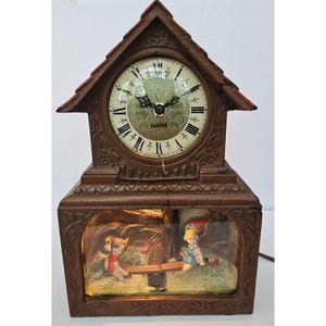 May include: A brown house-shaped clock with a gabled roof and a circular face featuring black Roman numerals and the text Warren. Below the clock face is an illuminated rectangular compartment containing two figures on a seesaw in a garden scene.