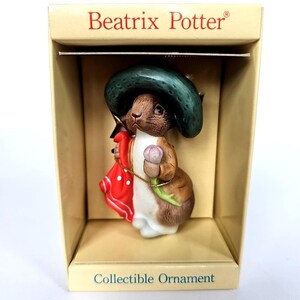 Puede incluir: Adorno coleccionable de un personaje de conejo con sombrero verde y chaqueta roja, sosteniendo una flor rosa. El adorno está en una caja con las palabras "Beatrix Potter" y "Collectible Ornament".