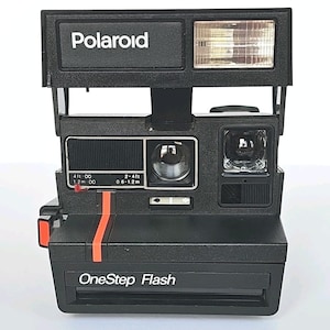 May include: Black Polaroid OneStep Flash instant camera with a built-in flash. The camera features a red stripe and the word "Polaroid" in white. It has a lens and a viewfinder. The text "OneStep Flash" is printed on the bottom.