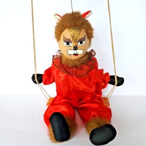 May include: A vintage marionette doll dressed in a red satin outfit with a fox-like face and brown fur. The doll is seated on a wooden swing, with black shoes and gloves. The doll has a painted face with detailed features.
