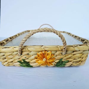 May include: A rectangular woven tray made of light tan natural fibers featuring two twisted rope handles on the sides. The front exterior is decorated with a single large orange flower with yellow highlights and two green leaves attached to the weave.