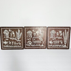 May include: Set of three square coasters with a dark brown background and white tribal art designs. Each coaster features a different scene with figures, huts, and plants. The coasters are likely made of a durable material.