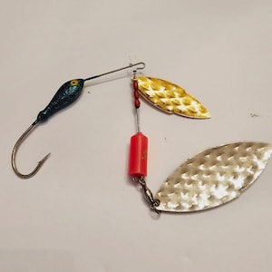 May include: Fishing lure with a blue glitter head and a large hook. The lure has two blades, one gold and one silver, and a red cylindrical body. The blades are textured and designed to attract fish.