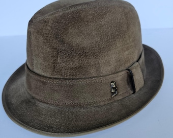 May include: A brown fedora hat with a suede-like texture. The hat features a dark band around the crown with a small decorative metal emblem. The brim is slightly upturned, and the hat has a classic, vintage style.