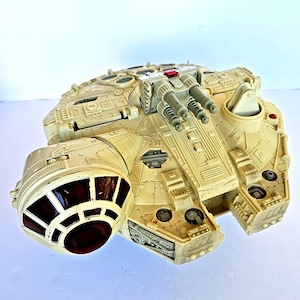 Può includere: Un'astronave giocattolo color beige con pannelli dettagliati, torrette e una luce rossa. Il giocattolo presenta un abitacolo circolare con finestre rosso scuro ed è progettato con molteplici componenti rettangolari e circolari.