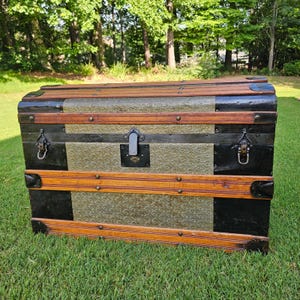 May include: A rectangular storage trunk featuring a domed lid, constructed with black metal panels, horizontal wooden slats, and embossed gold-toned metal sheets with a circular pattern. It includes dark metal corner reinforcements and a central latch.
