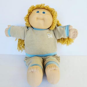 May include: A doll with yarn-like blonde hair, blue eyes, and a round face. It wears a beige long-sleeved shirt with a small white cat graphic and light blue trim, paired with matching beige trousers featuring light blue cuffs at the ankles.