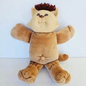 Puede incluir: Una muñeca de peluche con forma de gato, con cara de tela color canela, pelo de lana marrón y un cuerpo de terciopelo marrón suave. La muñeca presenta detalles faciales bordados, incluyendo ojos marrones y una nariz pequeña, con brazos extendidos.