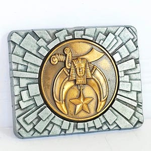 May include: A rectangular belt buckle featuring a central gold-toned circular emblem with a pharaoh head, two curved scimitar swords, and a five-pointed star. The surrounding border is a silver-toned textured geometric pattern of radiating blocks.
