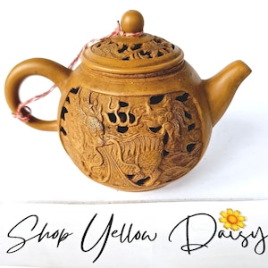 May include: A tan-colored teapot with intricate carvings of a dragon and floral patterns. The lid and body feature cut-out designs. The teapot has a handle, a spout, and a small knob on the lid. The text "Shop Yellow Daisy" is at the bottom.
