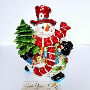 Puede incluir: Figura de muñeco de nieve de cerámica con sombrero de copa, bufanda y mitones rojos, sosteniendo un árbol de Navidad verde. El muñeco de nieve tiene una nariz de zanahoria y una sonrisa alegre. Con logo Shop Yellow Daisy.