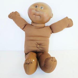 May include: A brown fabric doll with a smooth, rounded vinyl head, brown eyes, and a soft stuffed body. The doll features outstretched arms, a small belly button detail, and a solid brown complexion throughout its head and limbs.