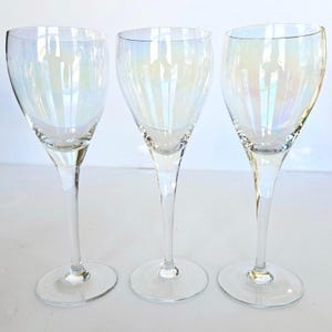 May include: Three clear iridescent wine glasses with tall stems and round bases. The glasses have a pearlescent sheen, reflecting light with a rainbow effect. The glasses are empty and appear to be made of glass.