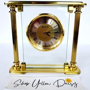 May include: A gold-toned quartz clock with a clear glass face. The clock face is gold with black Roman numerals and the brand name "Benchmark". The clock has a rectangular shape with gold pillars and a base.