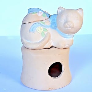 Puede incluir: Figura de gato de cerámica beige claro, con una cinta azul y detalles florales. El gato está posado sobre una base beige a juego con una abertura circular. La tapa del gato está decorada con rosas.