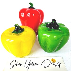May include: Three glass bell peppers in vibrant colors: red, yellow, and green. Each pepper has a dark green stem. The peppers are arranged on a white surface. The text "Shop Yellow Daisy" is at the bottom.