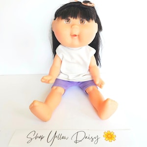 Könnte beinhalten: Eine Vintage-Puppe mit schwarzen Haaren, braunen Augen und einem runden Gesicht. Die Puppe trägt ein weißes T-Shirt und lila Shorts. Die Puppe sitzt mit ausgestreckten Beinen. Der Text "Shop Yellow Daisy" steht unten.