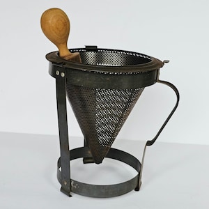Könnte beinhalten: Eine Passiermühle aus Metall mit einem konischen, perforierten Siebkorb, der von einem dunklen Metallrahmen mit Griff gehalten wird. Ein heller Holzstößel befindet sich im Sieb. Die Konstruktion verfügt über einen runden Standfuß.