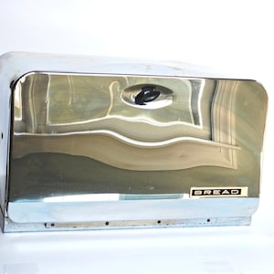 Op de afbeelding: Een vintage chromen broodtrommel met een gebogen bovenkant en een zwarte handgreep. Het woord "BREAD" staat op een rechthoekig label aan de voorkant. De trommel heeft een reflecterend oppervlak en de binnenkant is niet zichtbaar.