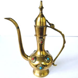 May include: A vintage brass decorative pitcher with a long spout and ornate handle. The pitcher features decorative red and turquoise accents. The design is reminiscent of Middle Eastern or Indian craftsmanship. The pitcher is likely used for serving beverages or as a decorative item.