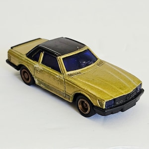 May include: A die-cast toy car featuring a metallic gold body, a black roof, and dark blue tinted windows. The vehicle has black bumpers, black wheels, and a detailed front grille. The paint shows signs of wear, and the design includes molded side vents and a sleek, low-profile coupe silhouette.