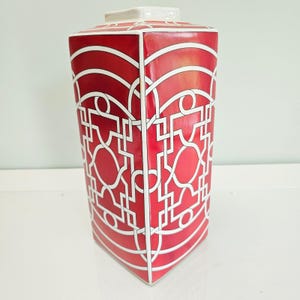 Peut inclure: Un vase rectangulaire haut en céramique présentant une couleur de base rouge vif avec un motif géométrique blanc composé de lignes et de cercles entrelacés. Le vase a une finition lisse et brillante et une ouverture carrée sur le dessus.
