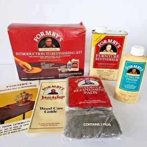 Puede incluir: Una colección de productos para el cuidado de la madera de Formby's, que incluye una caja roja con la etiqueta "Introduction to Refinishing Kit", una lata de "Furniture Refinisher" y una botella de "Tung Oil Finish". También se incluyen almohadillas de lana de acero y guías de cuidado de la madera.
