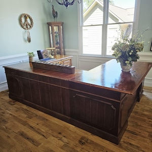Puede incluir: Un escritorio de madera grande en forma de L con acabado marrón oscuro y frente con paneles. La superficie del escritorio sostiene una computadora portátil, un portavelas de madera rectangular largo con aberturas circulares y una planta en maceta.