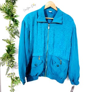 May include: A teal-colored jacket with a zip-up front and two front pockets. The upper portion of the jacket has a textured floral pattern. The jacket is hanging on a wooden hanger.