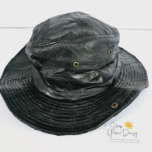 Op de afbeelding: Zwarte leren bucket hat met een brede rand en gestikte details. De hoed heeft verschillende kleine metalen oogjes en een textuur. Een stijlvol accessoire voor bescherming tegen de zon.