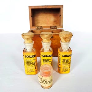May include: Three glass bottles with yellow labels labeled SCHWERT, each topped with a tan paper stopper, arranged in front of a small wooden box. A smaller, cylindrical plastic container labeled TESTING SALTS sits in the center foreground.