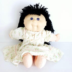 May include: A vintage doll with a round face, blue eyes, and black yarn hair. The doll wears a cream-colored dress with a floral pattern and pink trim. The doll's arms and legs are extended, and it is sitting.
