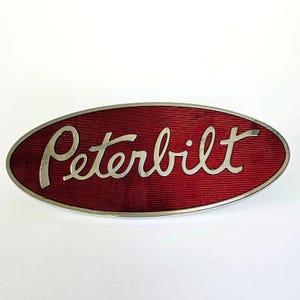May include: Oval-shaped emblem with the word "Peterbilt" in silver script lettering. The background is a deep red with a textured, ribbed pattern. The emblem has a silver border and is set against a white background.