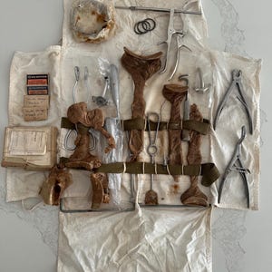 Puede incluir: Un kit médico vintage con varios instrumentos quirúrgicos. El kit incluye herramientas de metal, componentes de cuero marrón y pequeños paquetes de papel, todo dispuesto sobre una tela color crema. Los instrumentos están asegurados con correas verde oliva.