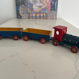 May include: A wooden toy train. The engine is red and dark blue, with yellow details. Two blue carriages with yellow interiors and red wheels are attached. The toy train is on a white surface. The train is approximately 30 cm long.